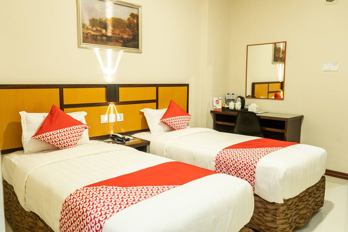A hotel room with two beds, each with white bedding and red accents, a nightstand with a phone, a desk with a mirror, and a painting on the wall.