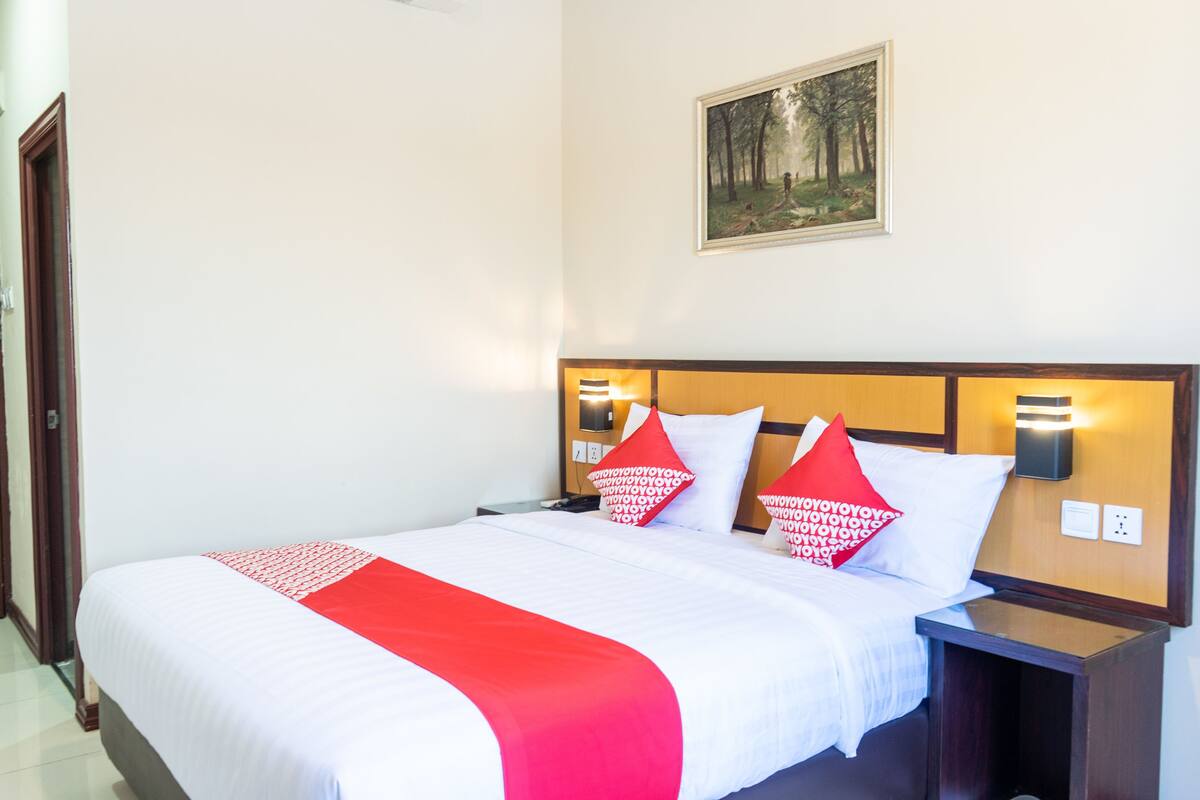 A neatly made bed with white and red bedding, a wooden headboard, and a painting on the wall above.