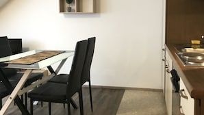 Une cuisine moderne avec un plan de travail en bois et un four encastré. On y trouve un coin repas avec une table en verre et des chaises noires. Une étagère en bois, ornée d’objets décoratifs, est fixée au mur.