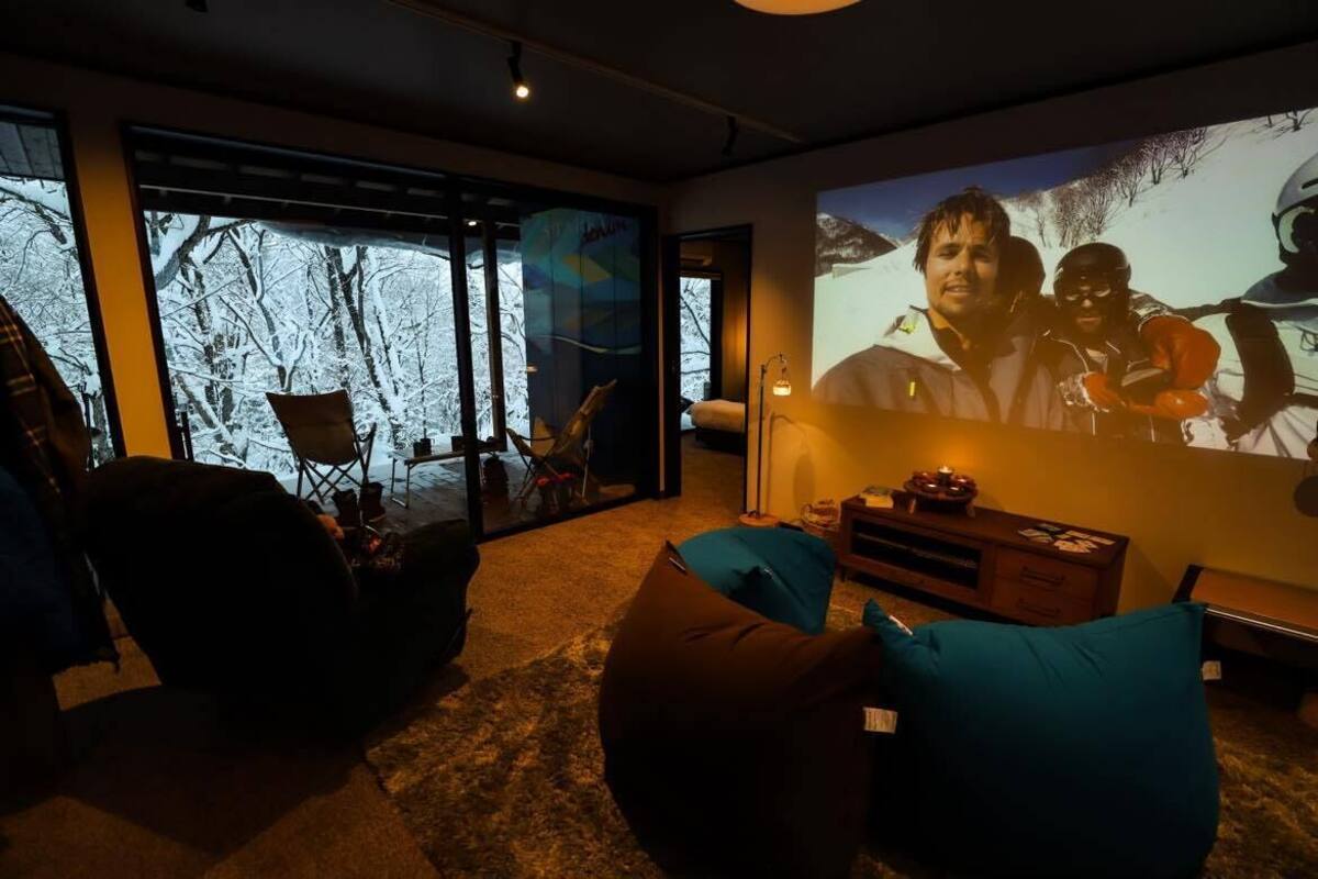 A cozy living room with a large screen displaying a skiing scene, comfortable seating, and a view of a snowy landscape through the glass door.