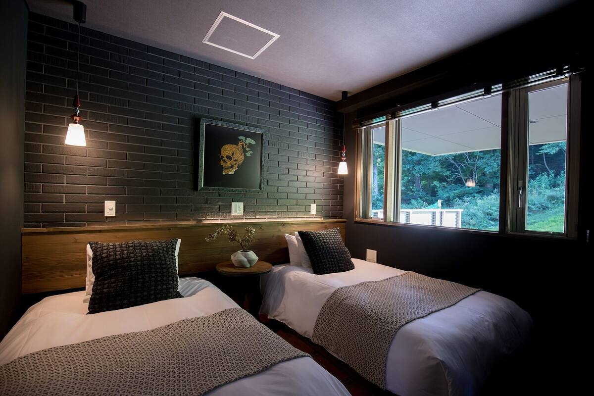 A room with two beds, a brick wall, a framed skull artwork, and a window with a view of greenery.