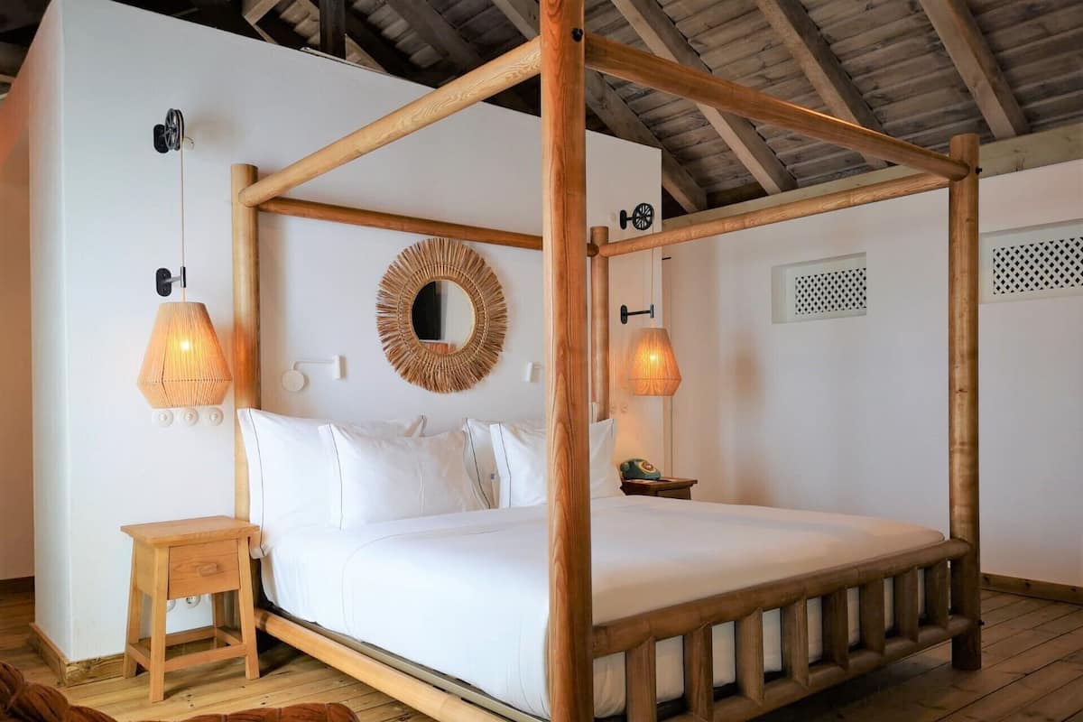 A four-poster bed with a wooden frame, two wall-mounted lamps, a small bedside table, and a woven wall decoration.