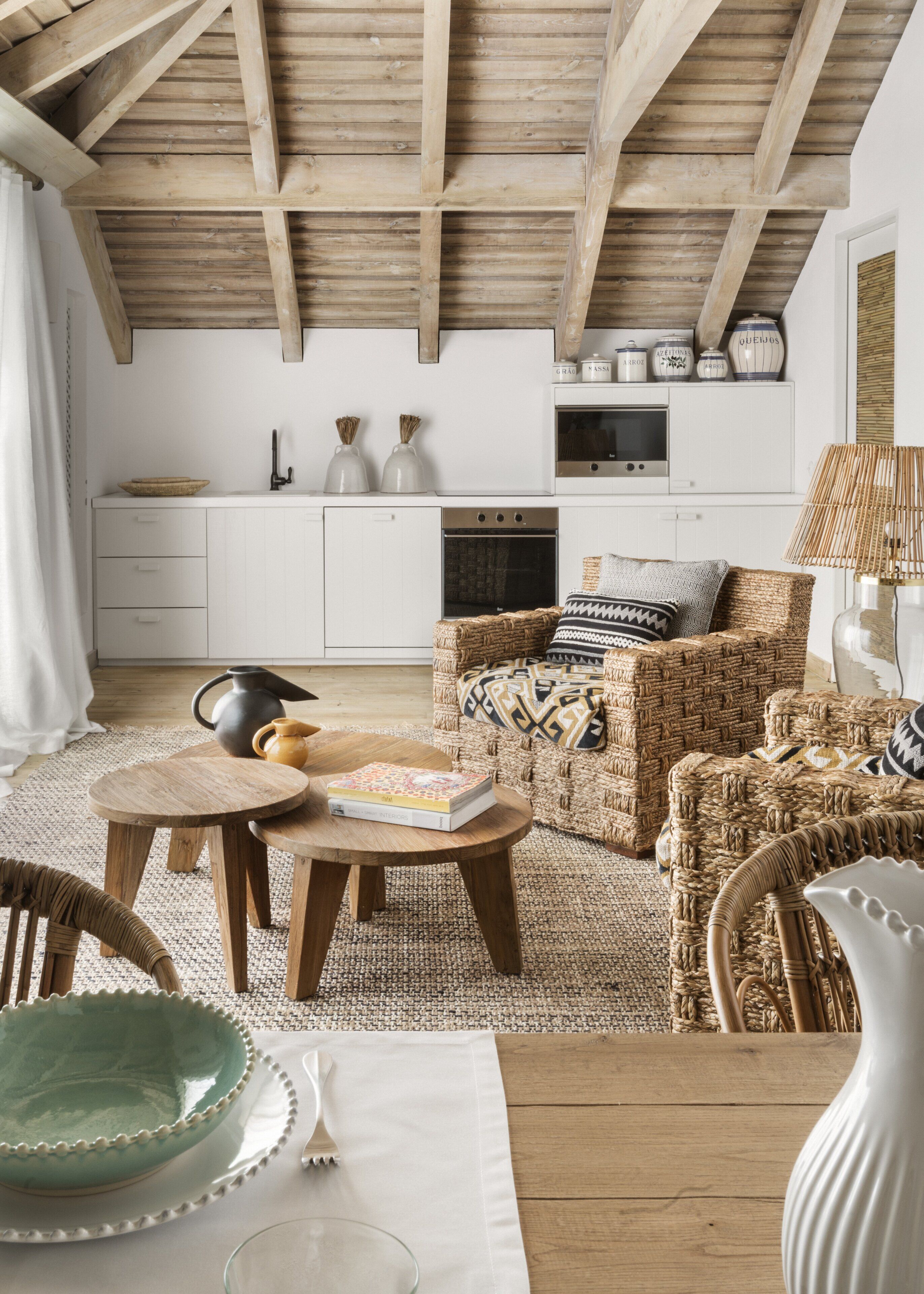 A cozy living room with a wooden ceiling, a wicker sofa, a dining table with a book, and a kitchen in the background.