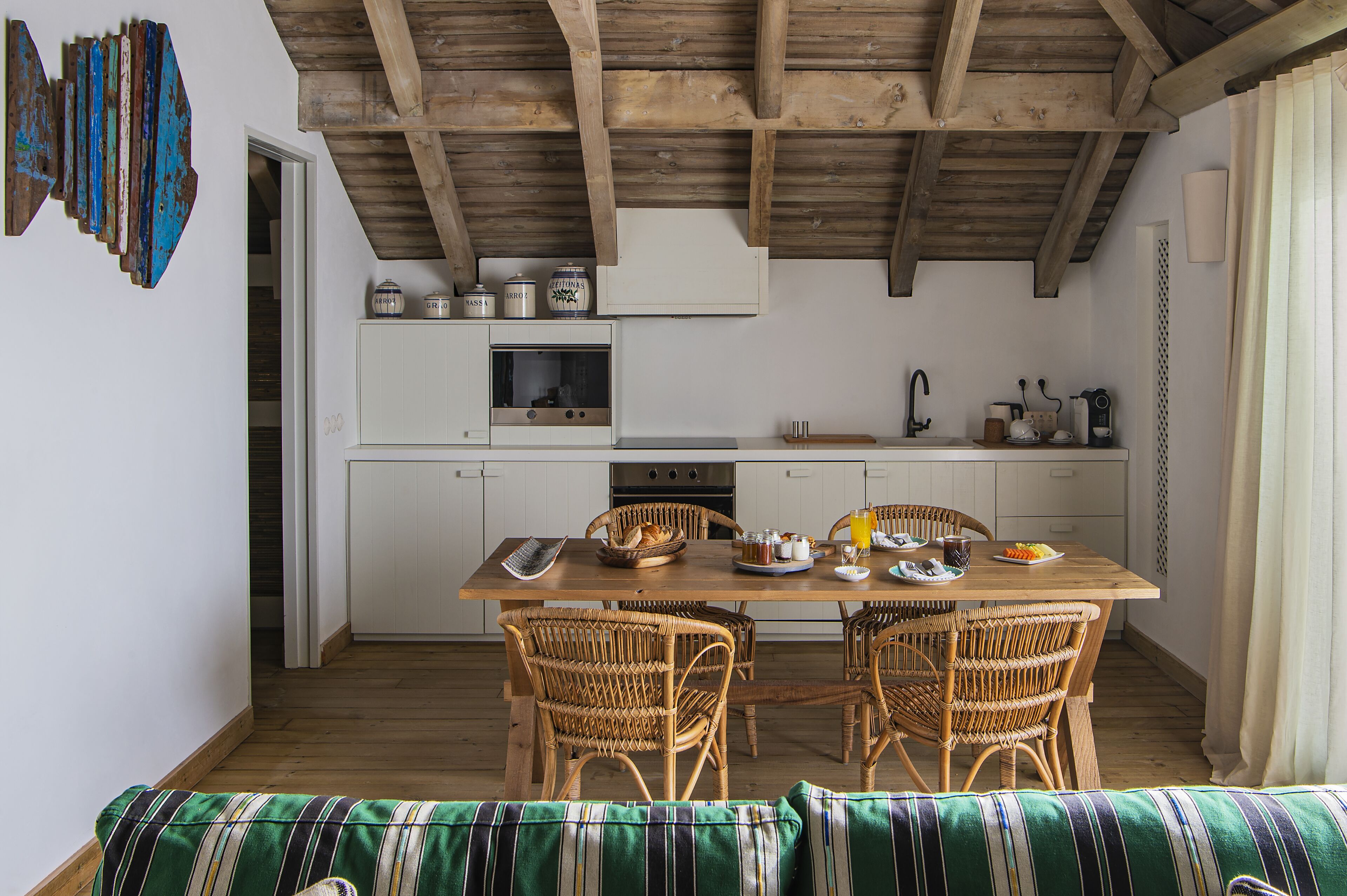 Une cuisine chaleureuse avec des poutres en bois, une table à manger avec des chaises et un canapé agrémenté d’un coussin à rayures.