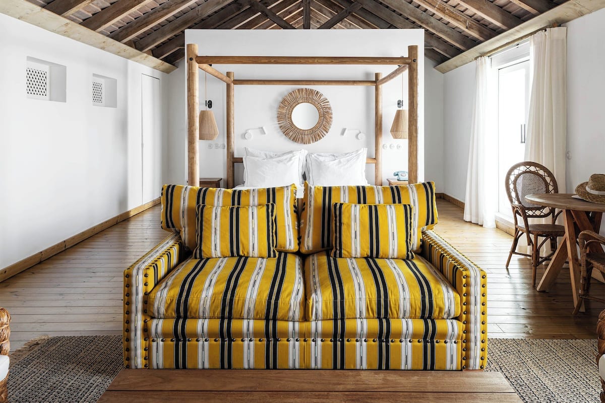 A room with a high wooden ceiling, a bed with a canopy, a dining area with a table and chairs, and a wicker chair.