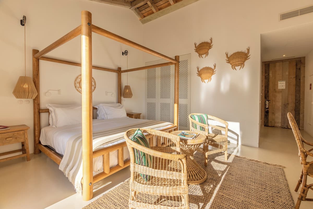 A four-poster bed with a wooden frame, two wicker chairs, a small table, and a woven rug.