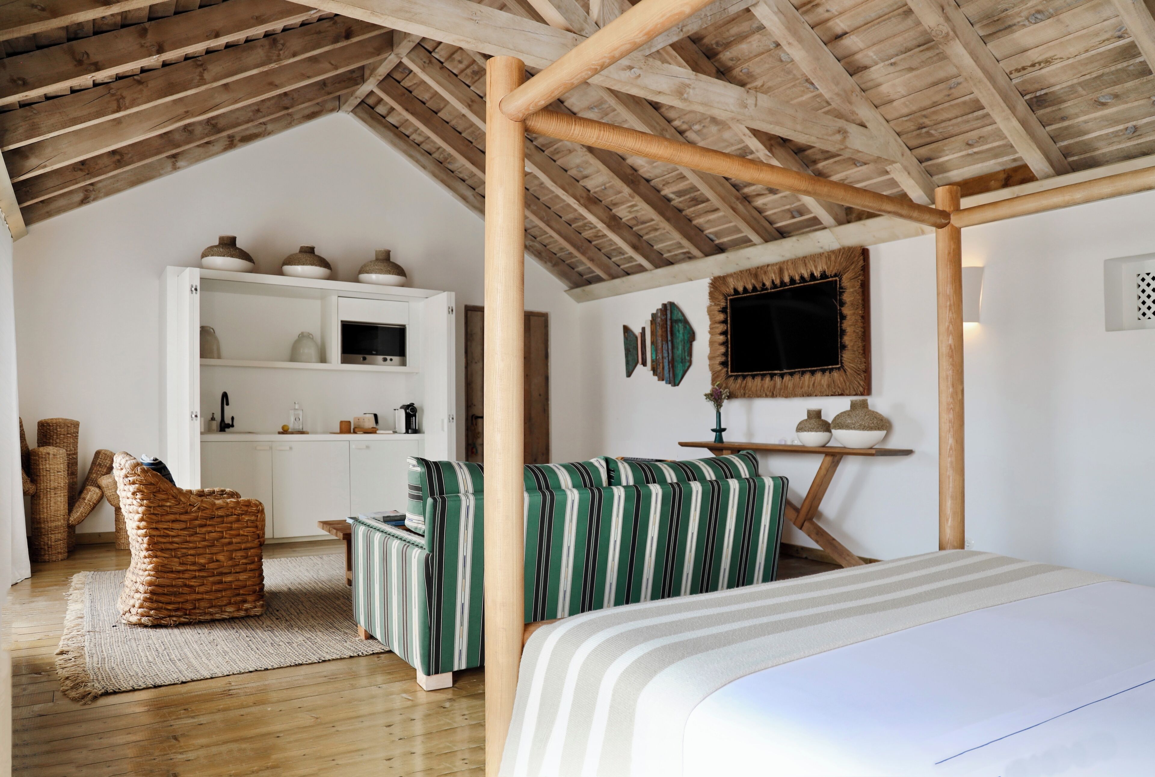A room with a high wooden ceiling, a bed, a striped sofa, a white cabinet, and a woven chair.