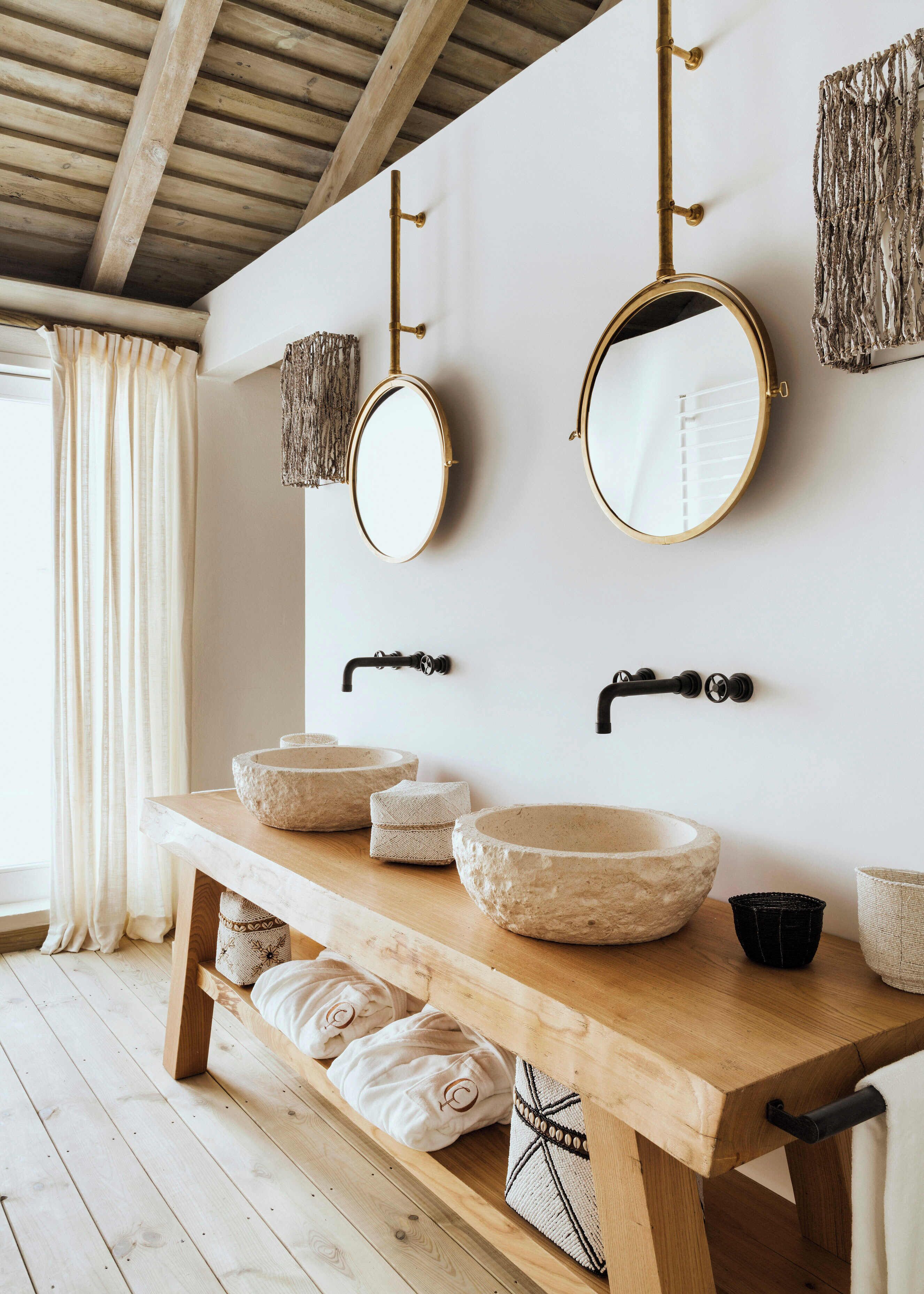 A bathroom with two round mirrors, a wooden vanity with three stone sinks, and a wooden bench with towels.