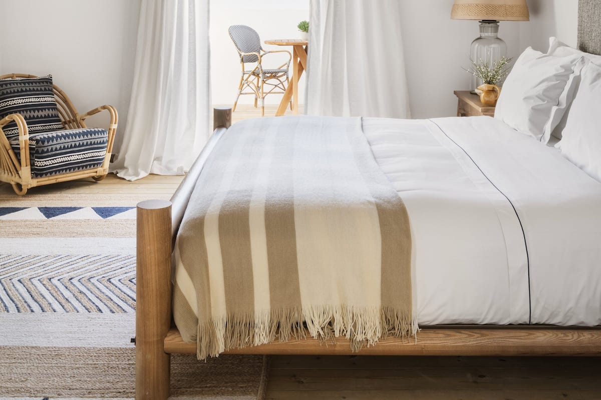 A bedroom with a wooden ceiling, a bed with a striped blanket, a bedside lamp, a chair, and a view of the outdoors.