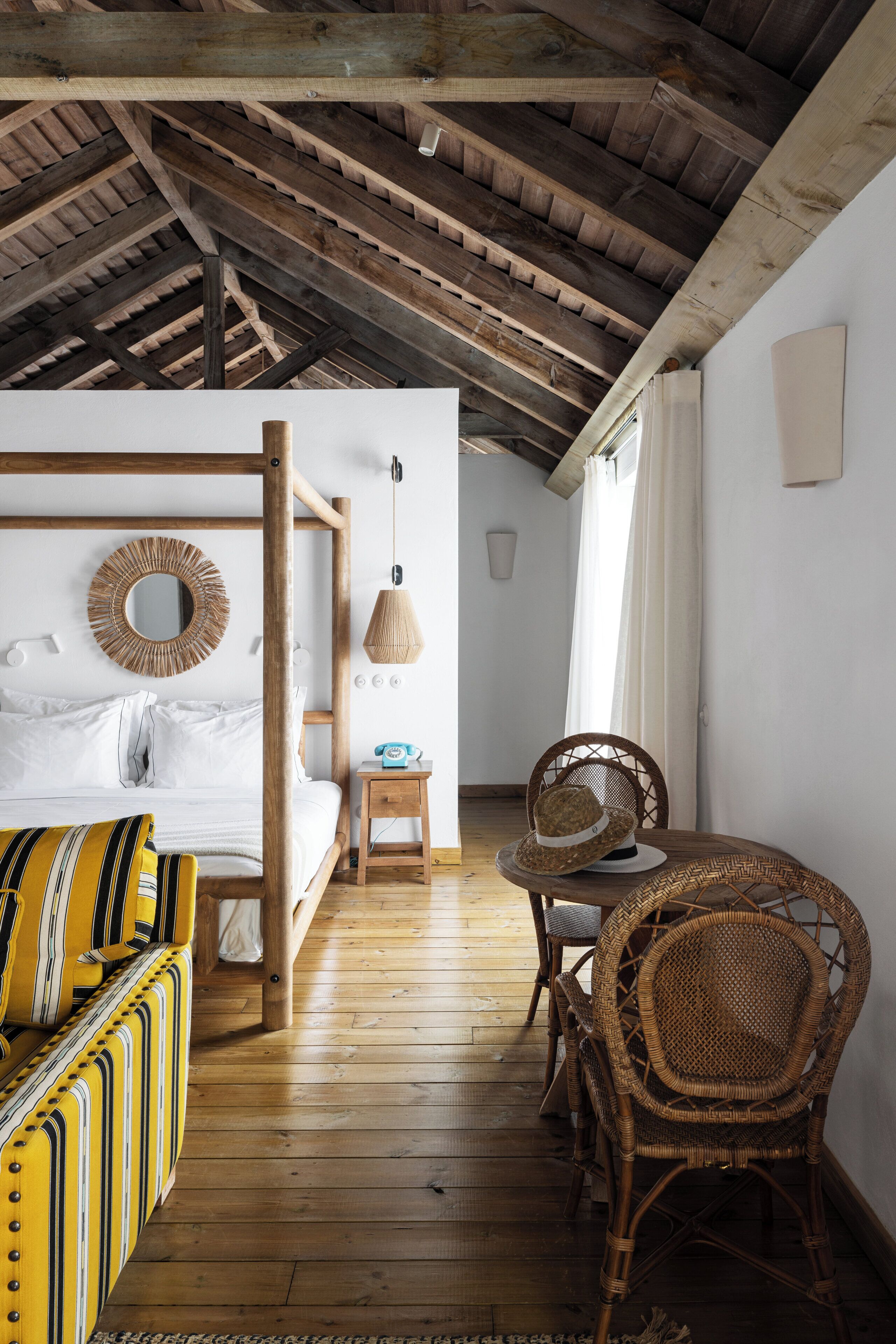 A room with a wooden ceiling, a bed with white bedding, a yellow-striped sofa, a wicker chair, and a small table with a lamp.
