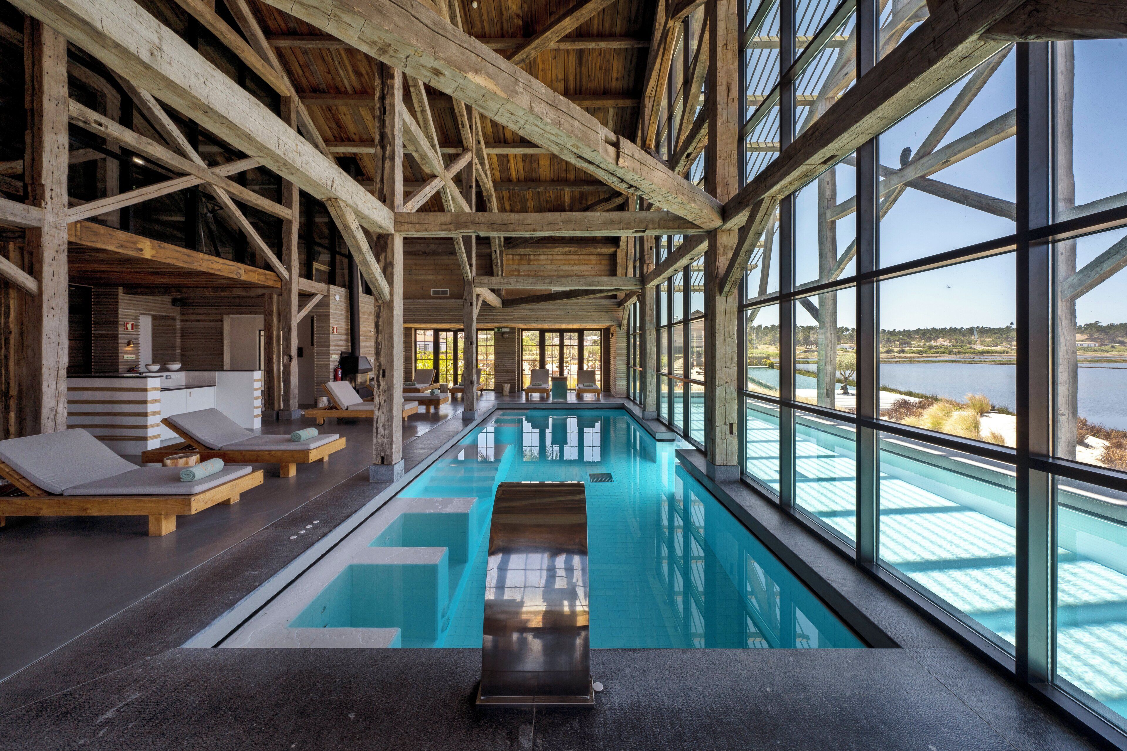 En indendørs swimmingpool med træbjælker, liggestole og udsigt til søen.