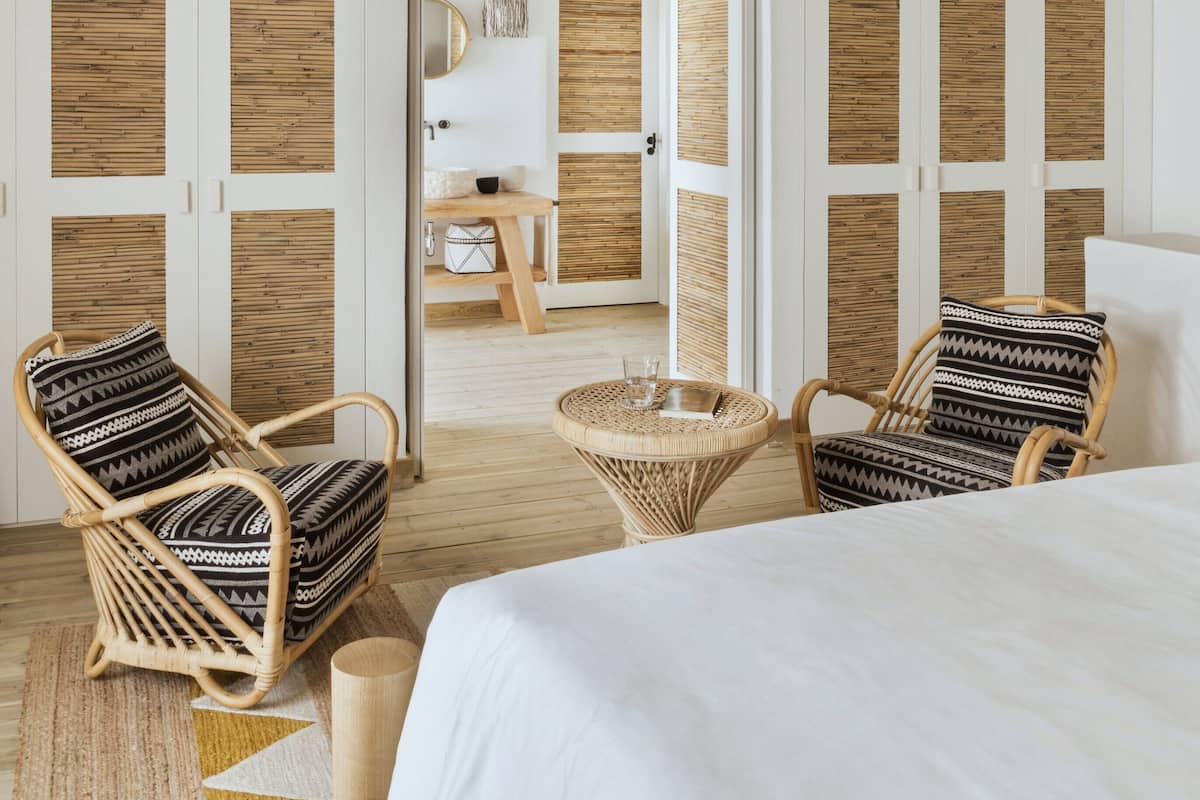 A spacious room with a high ceiling, wooden beams, and a large bed with white bedding. There are two wicker chairs with patterned cushions and a small round table with a glass.