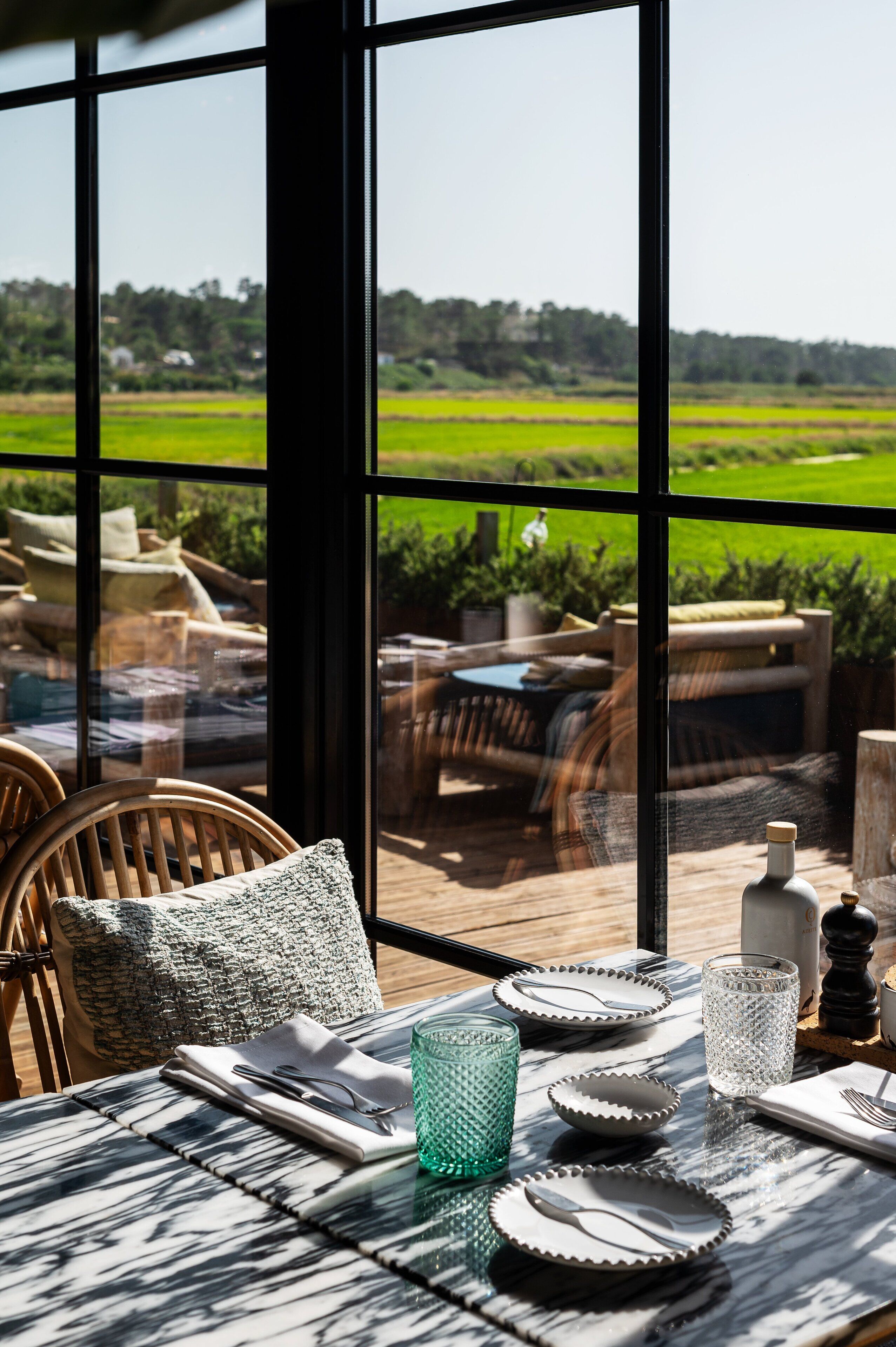 Un comedor con una mesa dispuesta para cuatro personas, vistas a un paisaje verde exuberante a través de amplios ventanales y una zona de estar exterior con mobiliario de mimbre.