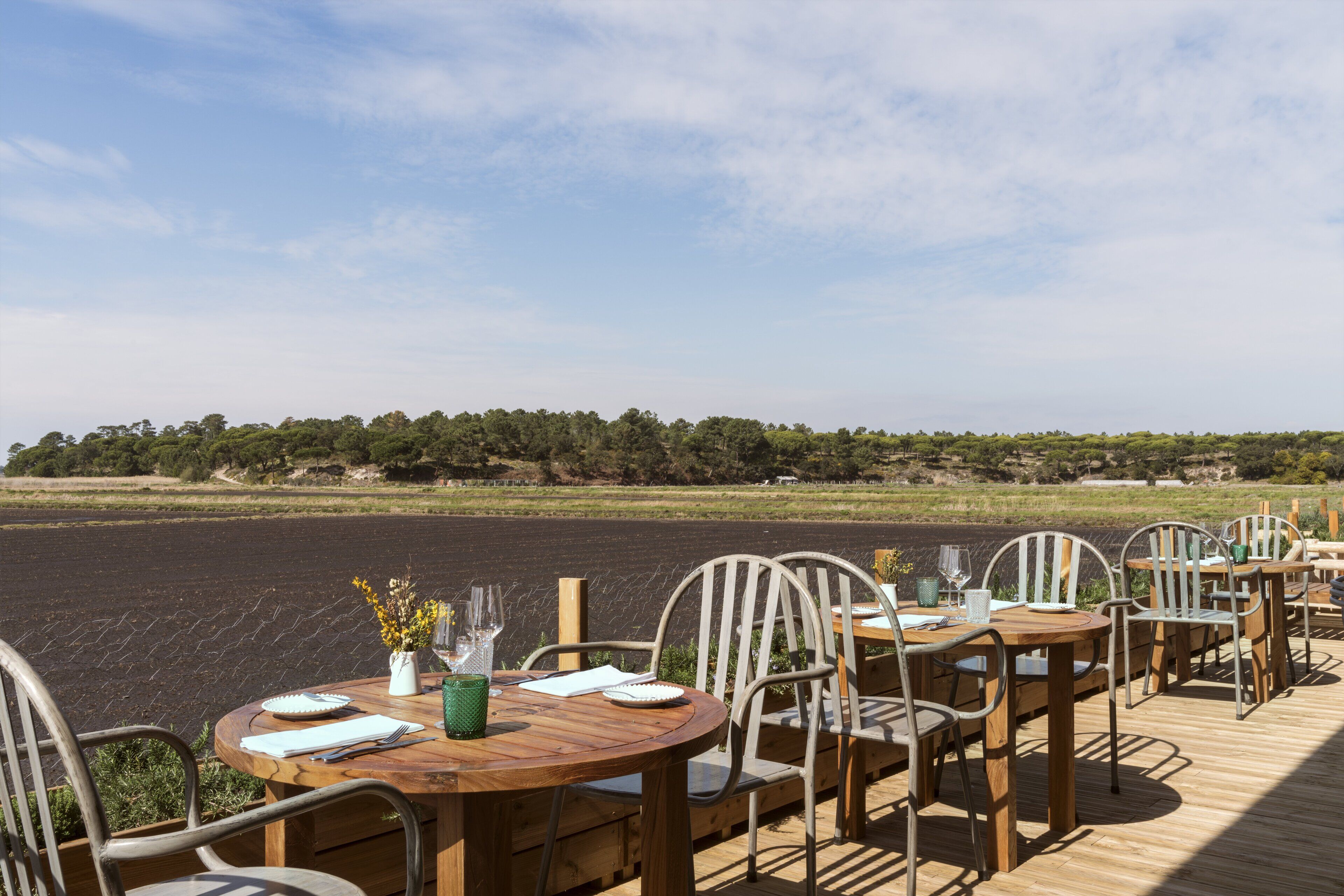 An outdoor dining area with wooden tables and chairs, overlooking a body of water and a distant tree line.