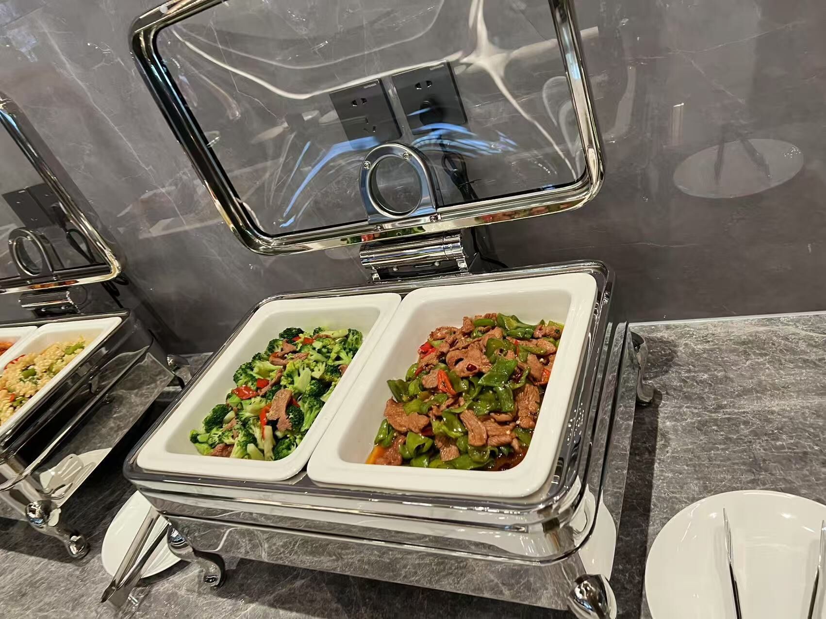 A buffet setup with two white chafing dishes containing stir-fried vegetables and meat, set on a marble countertop.