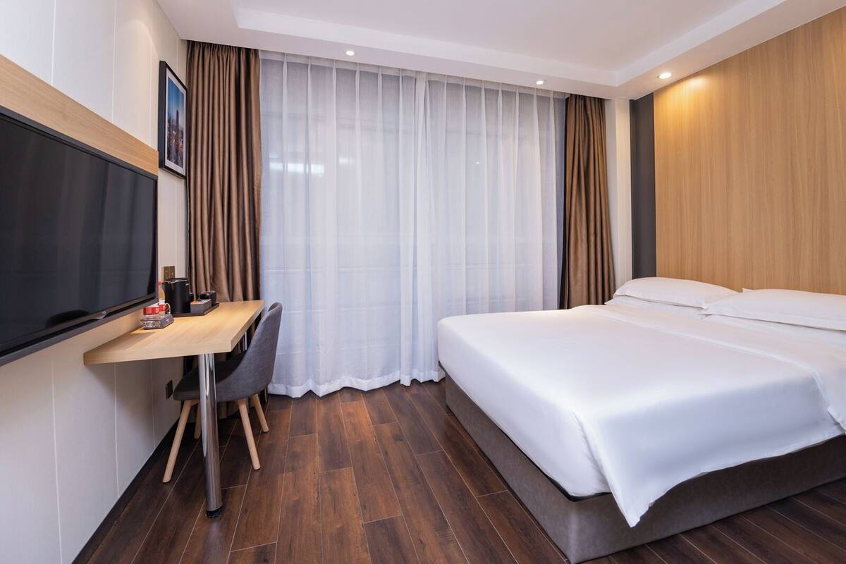 Habitación de hotel con cama, escritorio, silla, televisión y ventana con cortinas.