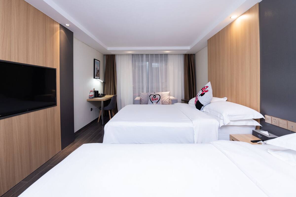 Habitación de hotel con dos camas, un escritorio con silla, un televisor de pantalla plana en la pared y una ventana con cortinas.