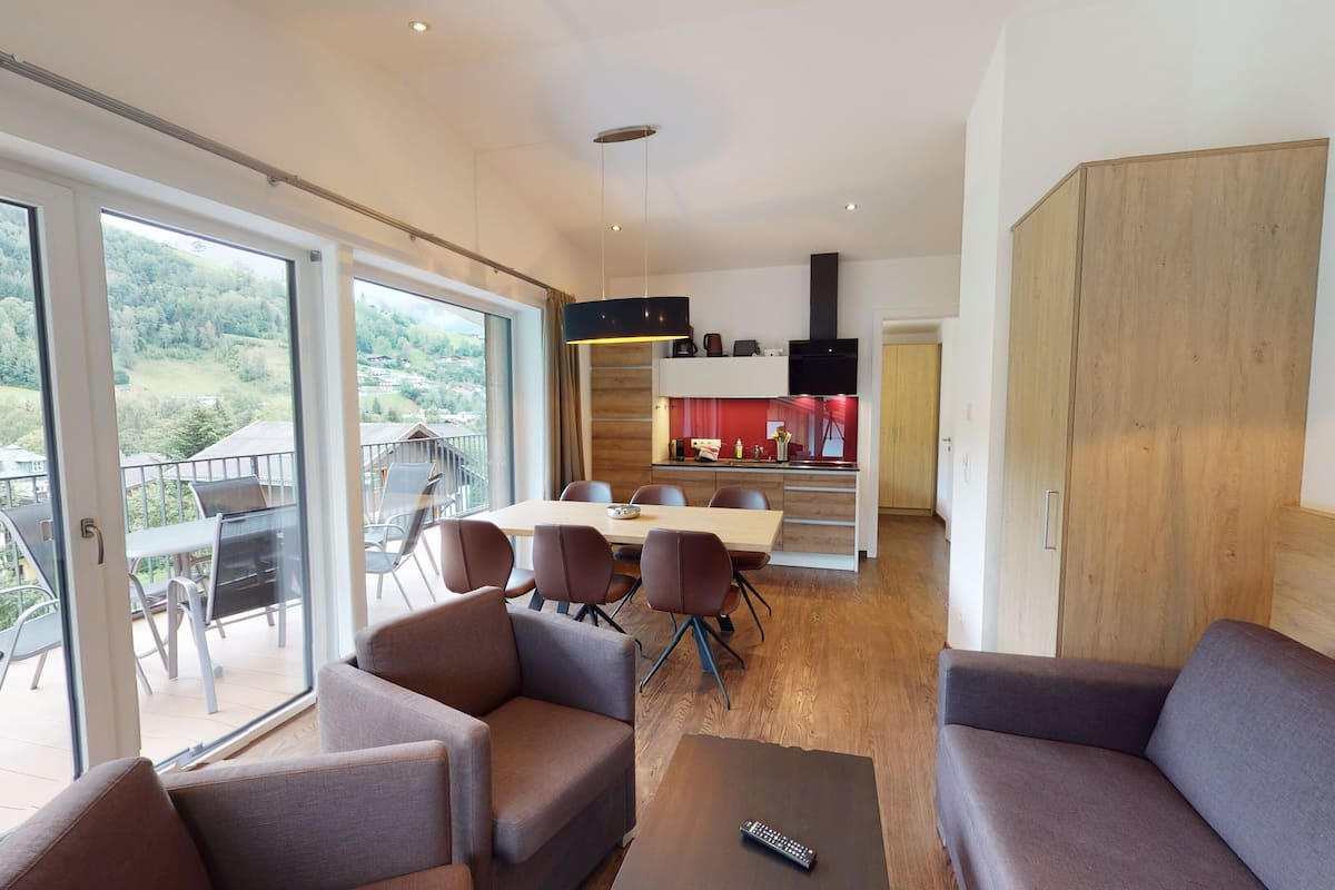 A modern living room with a large sliding glass door leading to a balcony, a dining area with a kitchen in the background, and a wooden wardrobe.