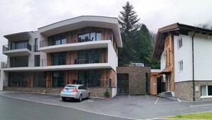 Un edificio moderno de varios pisos con balcones, un coche estacionado en la parte delantera y montañas al fondo.
