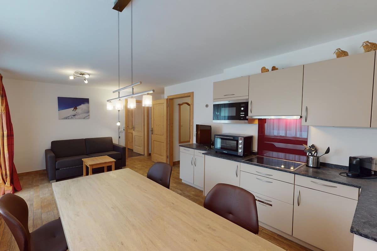 A modern kitchen with a microwave, toaster, and coffee maker. There is a dining area with a table and chairs. The room has wooden flooring and a large window with curtains.