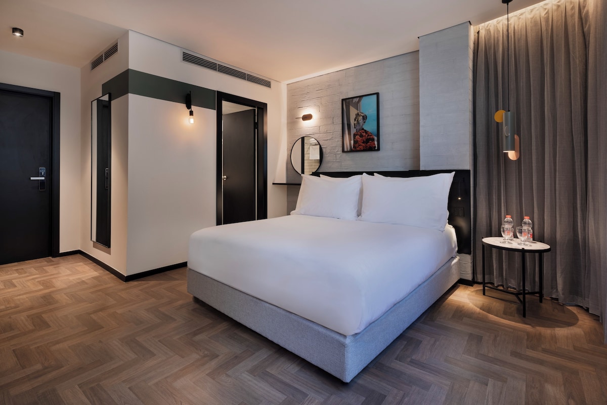 Ein modernes Hotelzimmer mit einem großen Bett, einem Nachttisch mit zwei Flaschen, einem Spiegel und einem Gemälde an der Wand.