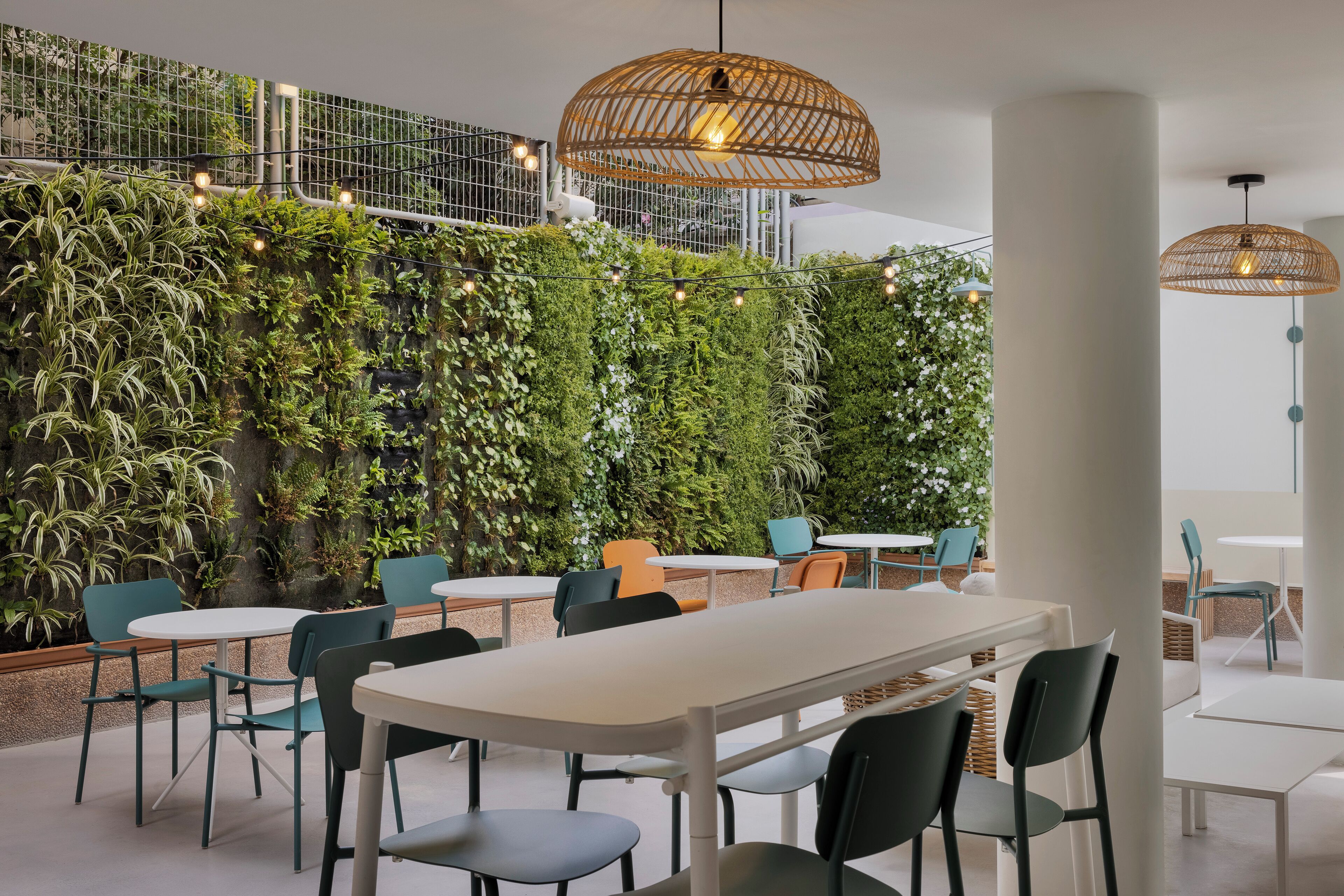 A modern indoor dining area with white tables and chairs, a green vertical garden wall, and wicker pendant lights.