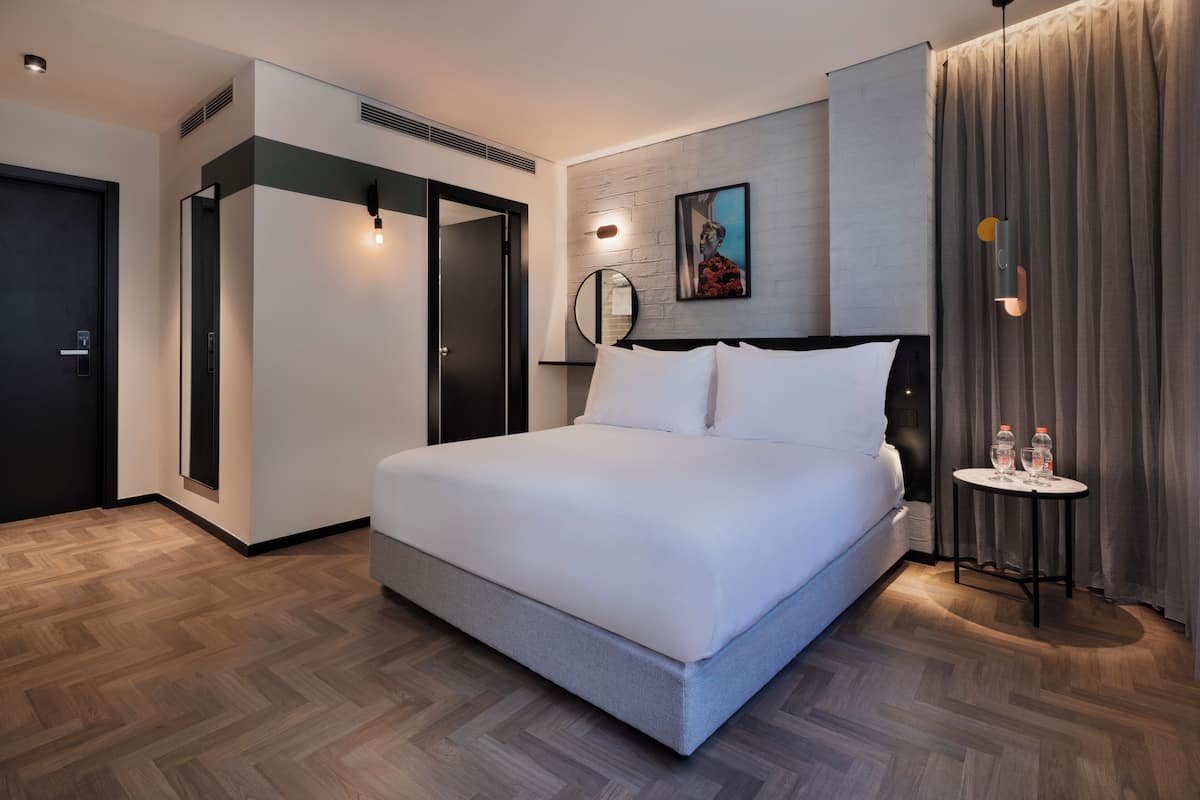 A modern hotel room with a large bed, a bedside table with two bottles, a mirror, and a painting on the wall.