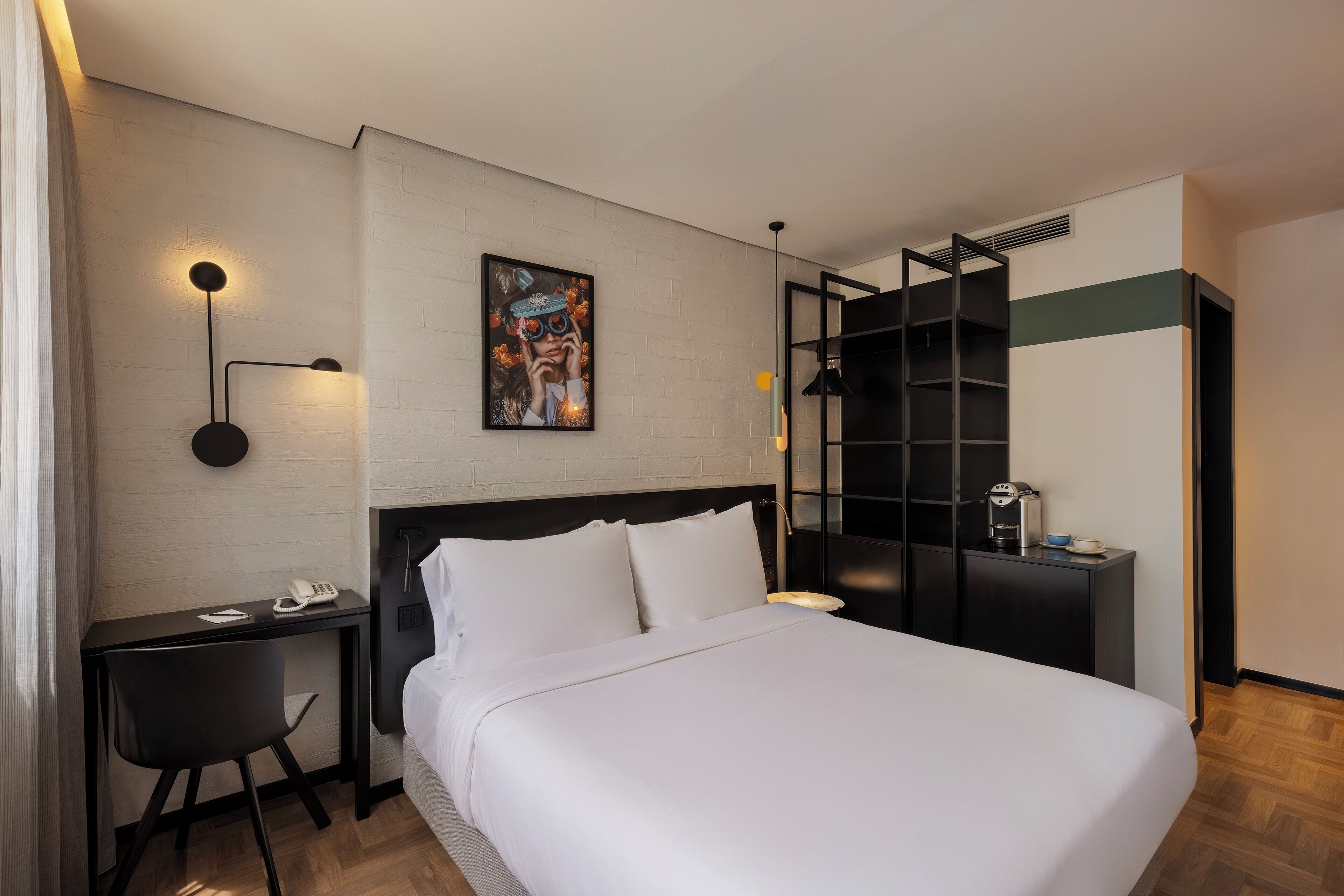 A hotel room with a large bed, a desk with a chair, a bookshelf, and a painting on the wall.