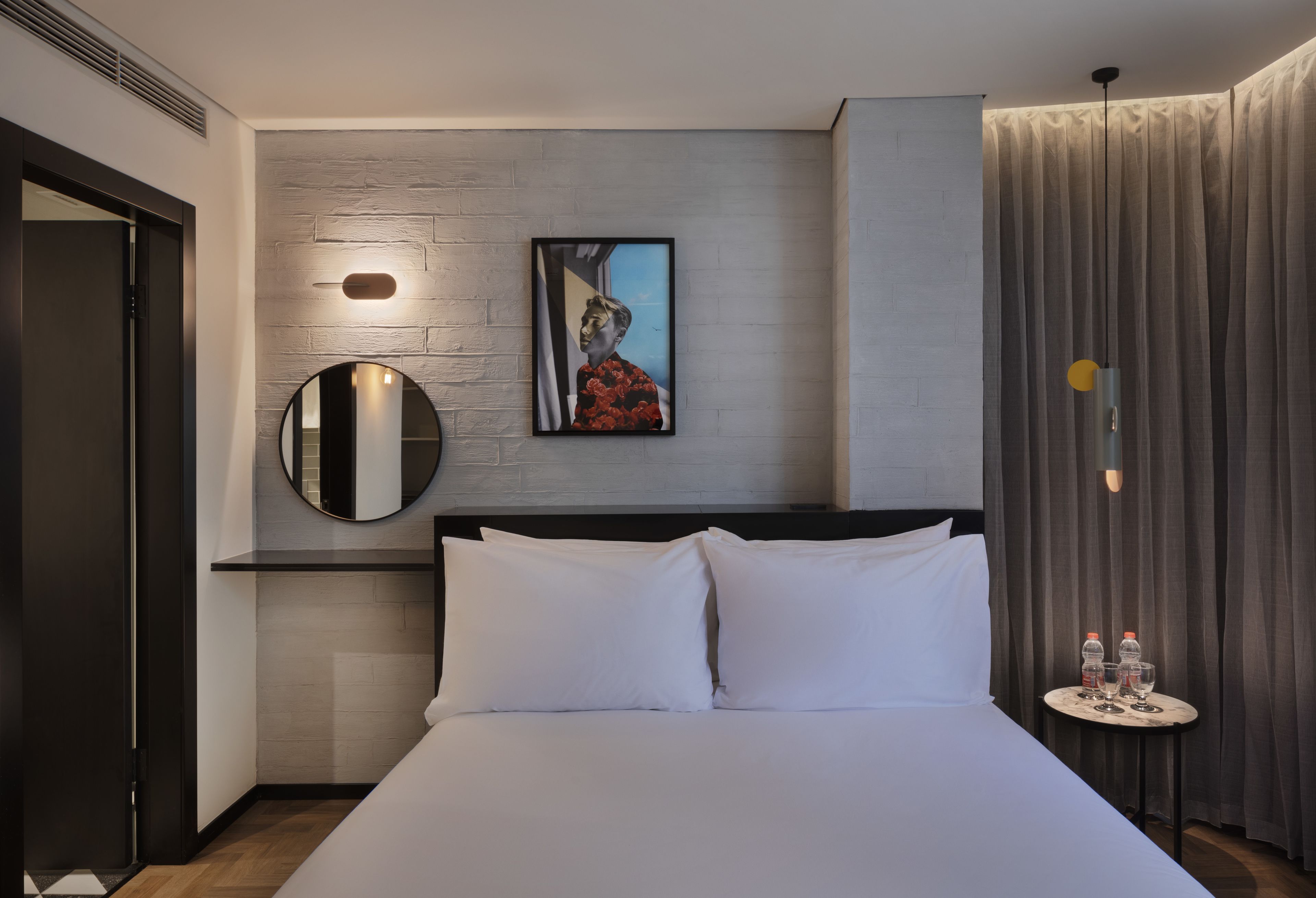 Una habitación de hotel moderna con una cama grande, un espejo redondo, una obra de arte enmarcada y una pequeña mesa con botellas de agua.