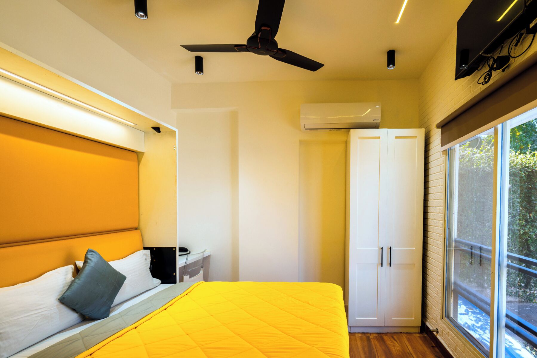 A bedroom with a bed, a ceiling fan, an air conditioning unit, a wardrobe, and a window with a view of greenery.