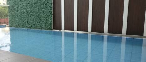 Una piscina con suelo de baldosas, rodeada por un seto verde y un muro de madera.