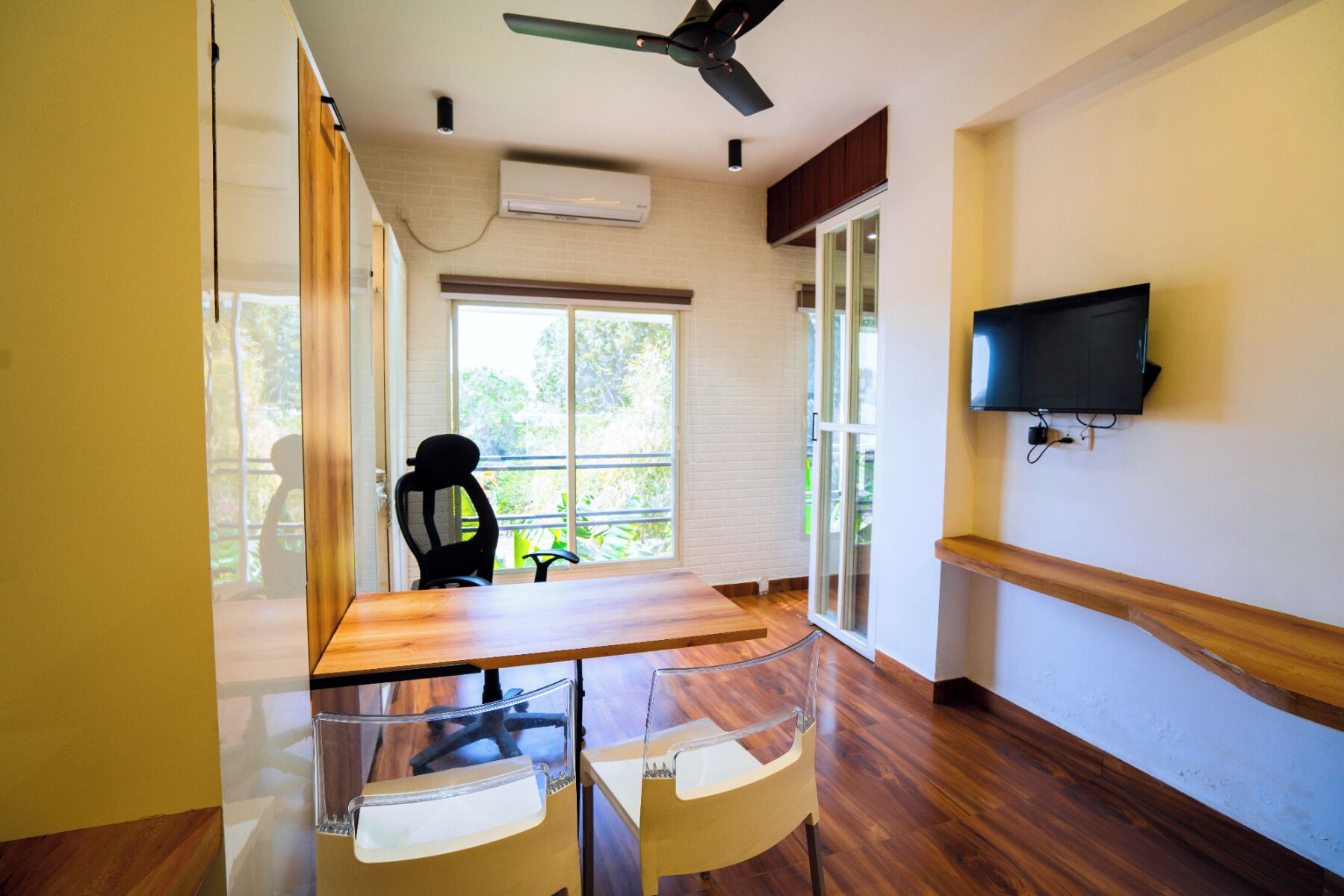 A room with a wooden table, chairs, a ceiling fan, a TV mounted on the wall, and a window with a view of greenery.