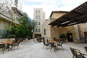 An outdoor cafe area with tables and chairs, a stone building, and a wooden pergola. - HI Agron Jerusalem Hostel (Jerusalem)