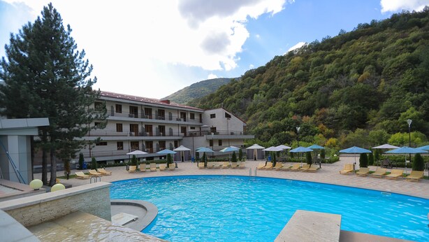 Una piscina con sillones de descanso y sombrillas, rodeada por un edificio con varios balcones y un paisaje montañoso de fondo.