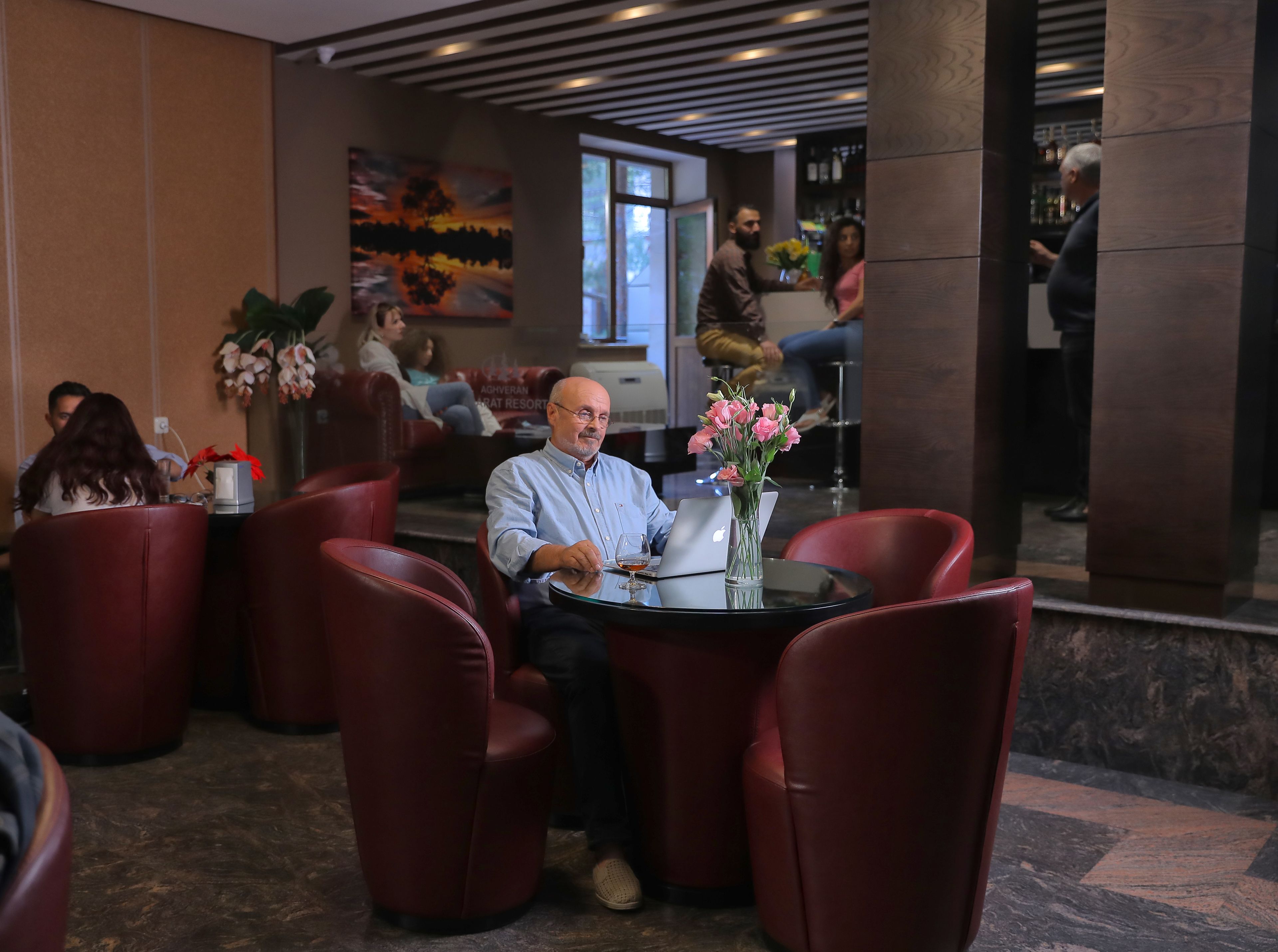 A man seated at a table with a laptop and a vase of flowers. Red leather chairs surround the table. In the background, there are people sitting and standing, and a bar area is visible.