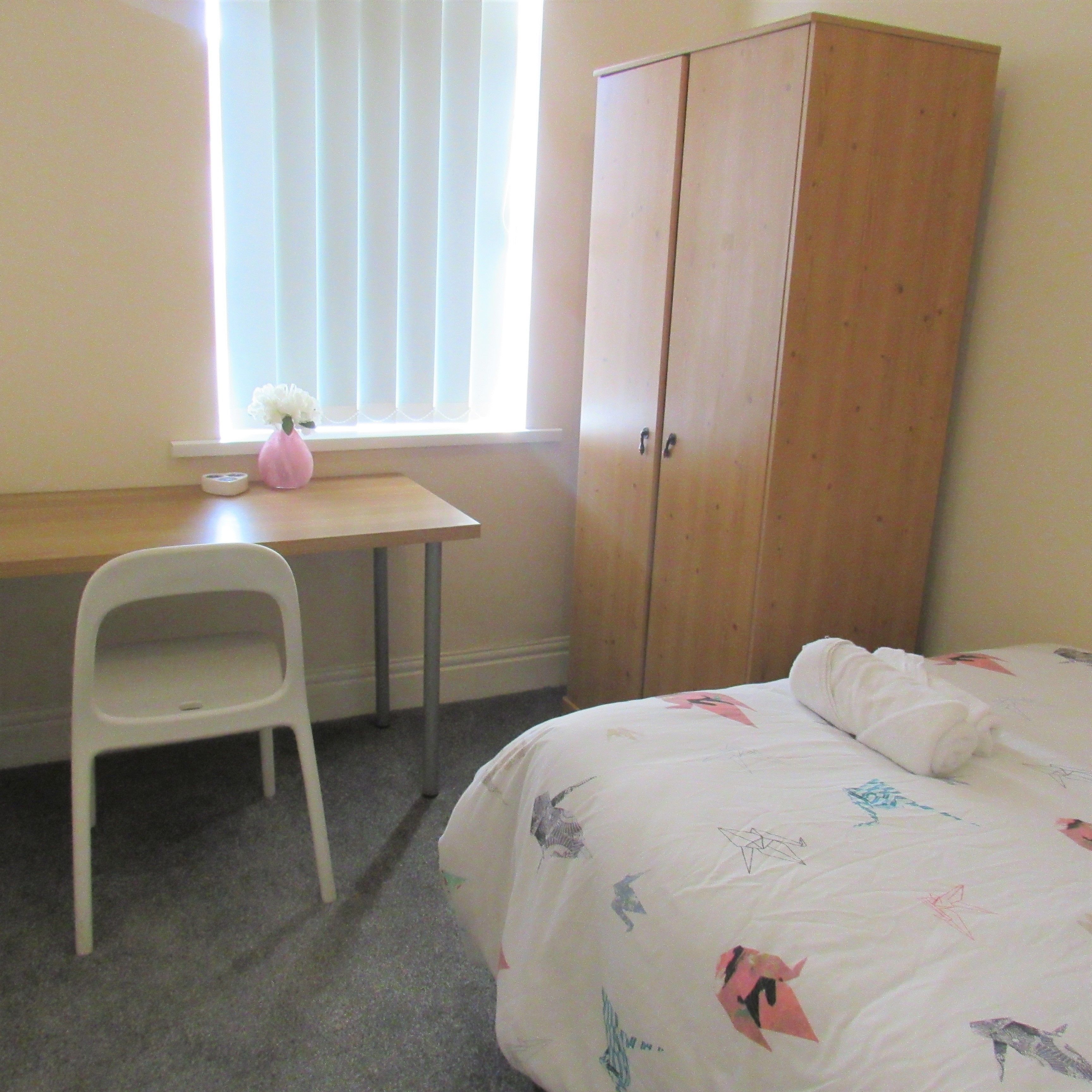 A bedroom with a bed, a desk, a chair, a wardrobe, and a window with blinds.