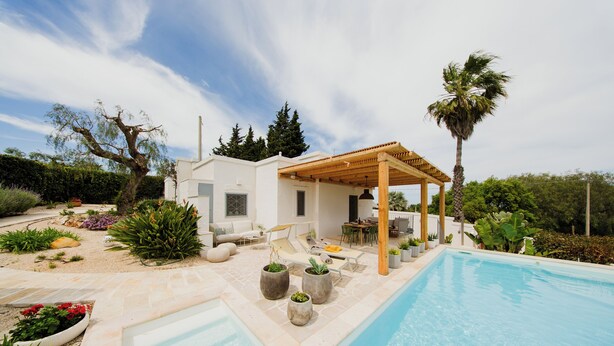 Un área junto a la piscina con un pérgola, mobiliario exterior y plantas en macetas.