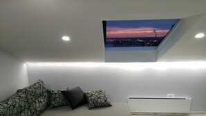Ein modernes Loft-Schlafzimmer mit grauem Sofa, gemusterten Kissen, einem kleinen Tisch und einem Dachfenster mit Blick auf eine Stadtlandschaft im Abendrot.