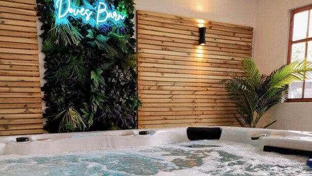 A hot tub area with a neon sign reading "Doves Barn," surrounded by wooden paneling and lush greenery. - Doves Barn (Ipswich)