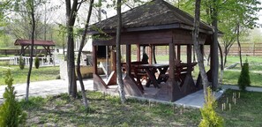 A wooden gazebo with a tiled floor and benches, surrounded by trees and a fenced area.