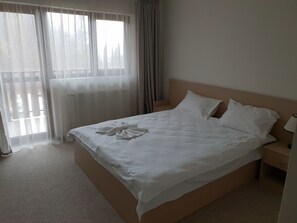 A neatly made bed with white linens and pillows, a wooden headboard, a bedside table with a lamp, and sheer curtains on a window.