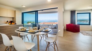 A modern dining area with a table set for four, large windows offering a view of the sea, and a red sofa in the adjacent living area. - JAZZ at Rova Apartment Sol (Malinska-Dubasnica)