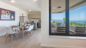 A modern dining area with a table set for four, a kitchen in the background, and a balcony with a view of the sea. - JAZZ at Rova Apartment Sol (Malinska-Dubasnica)