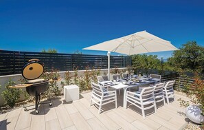 Eine Terrasse im Freien mit Grill, Esstisch, Stühlen und Sonnenschirm.