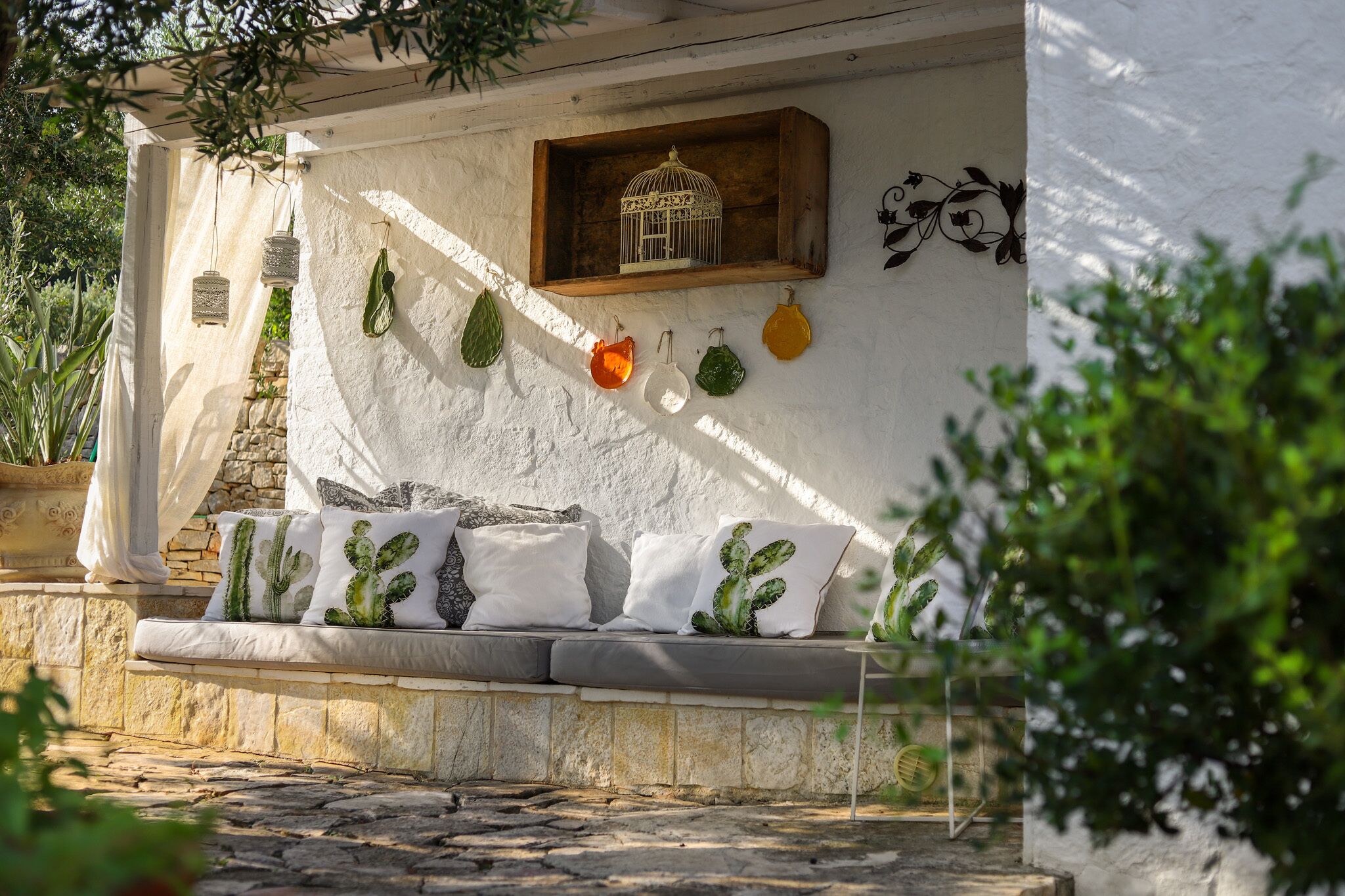 A stone bench with cactus-themed cushions, a wooden shelf with a birdcage, and hanging planters against a white wall.