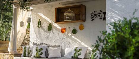 A stone bench with cactus-themed cushions, a wooden shelf with a birdcage, and hanging planters against a white wall.