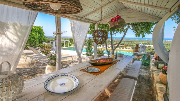 A wooden table with a white plate and a bowl of fruit, a wicker basket, and a hanging basket with flowers, overlooking a pool and a scenic landscape. - TD Casa Torchiano Hilltop Pool with Sea View (Monopoli)