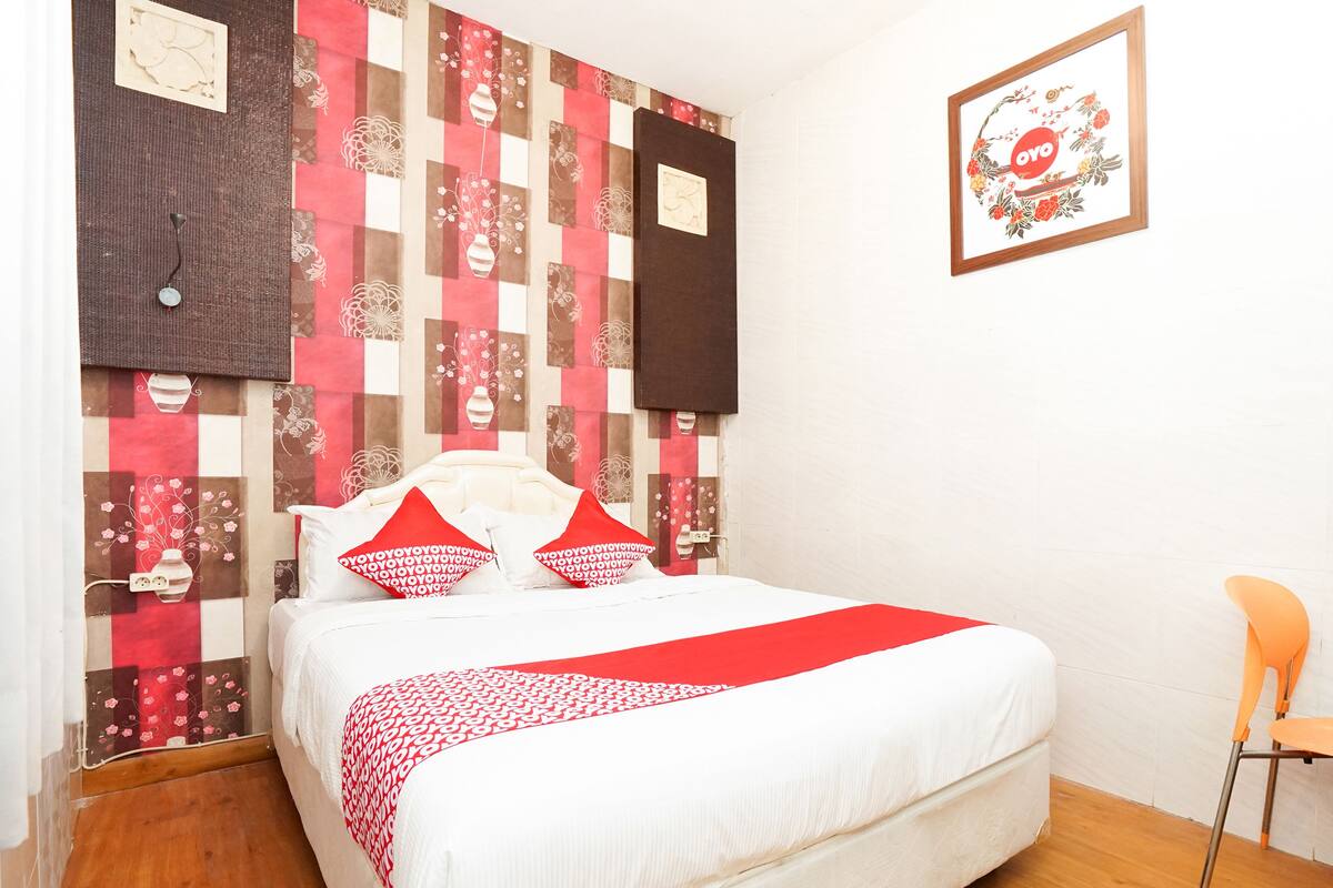 A bedroom with a bed, pillows, a red and white patterned blanket, a wooden floor, and a wall with a quilted design.