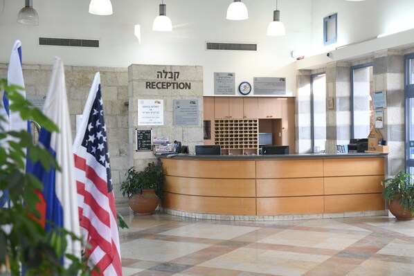 Zona de recepción con mostrador, banderas estadounidense e israelí, un reloj y señalización informativa.
