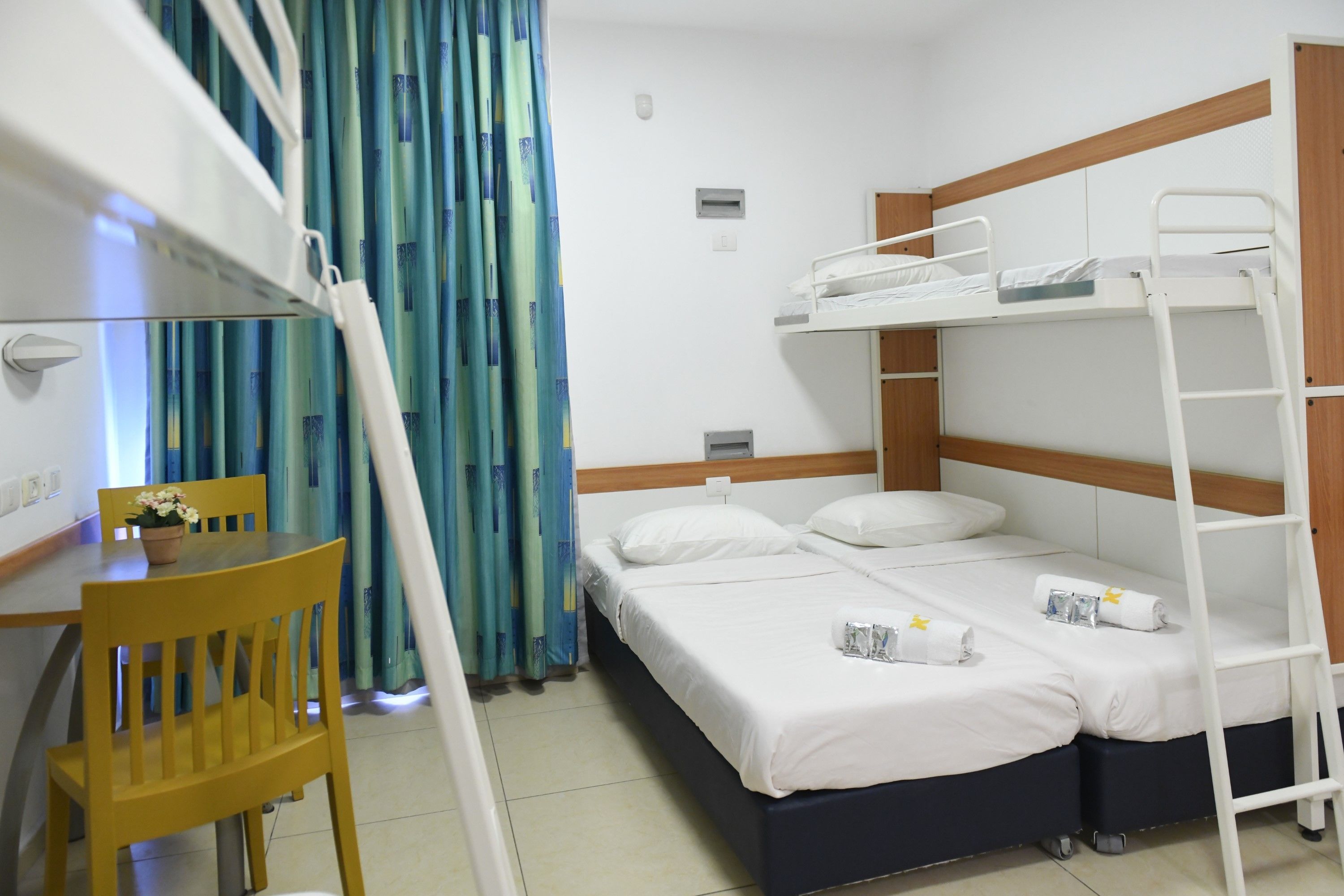A bunk bed room with a desk and chair, a small table with a flower, and a curtain with a blue and green pattern.